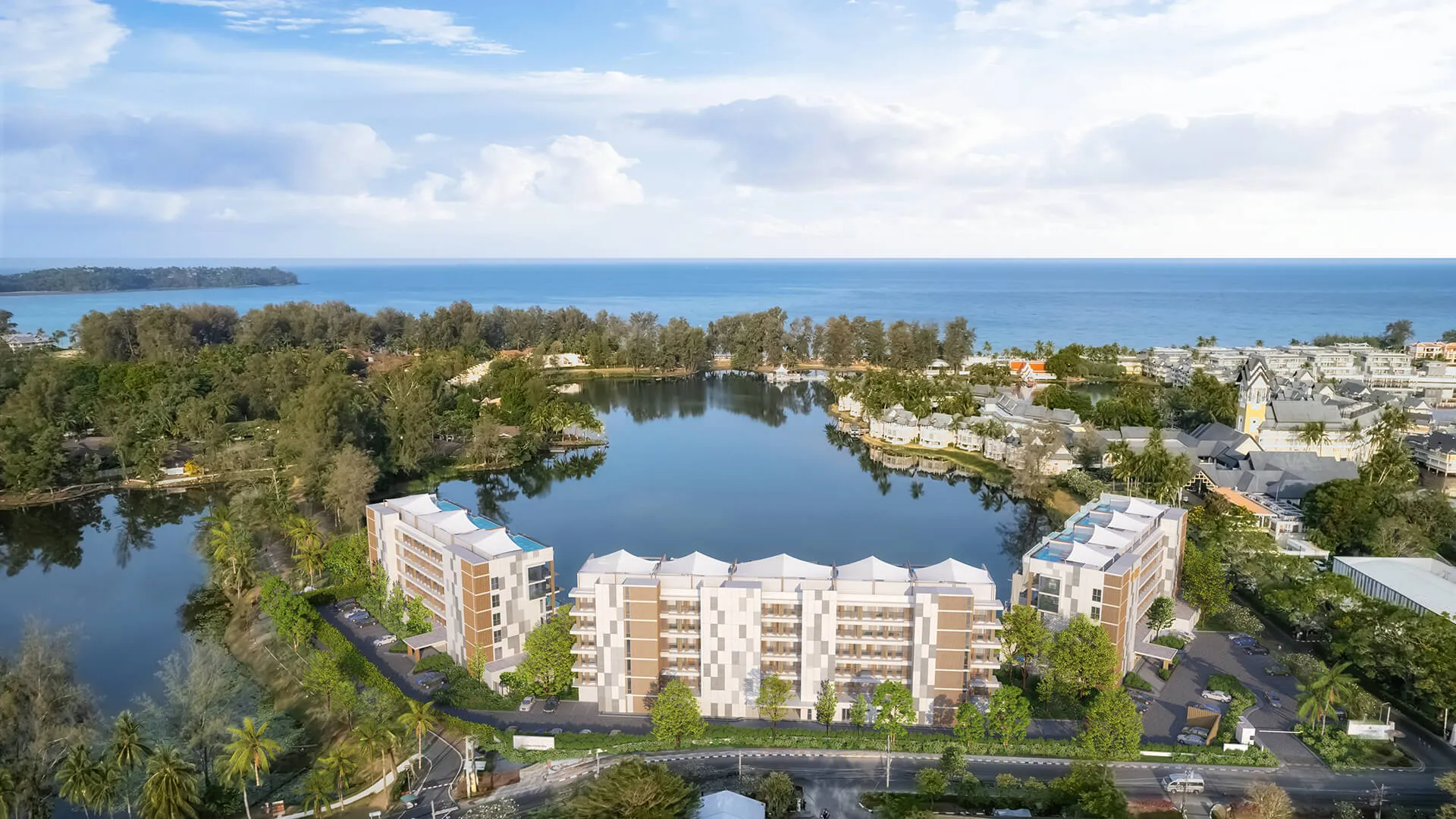 Angsana Oceanview Residences Phuket | A coastal aerial view showing buildings surrounding a calm central lake, with a lush tree line leading to a serene ocean.