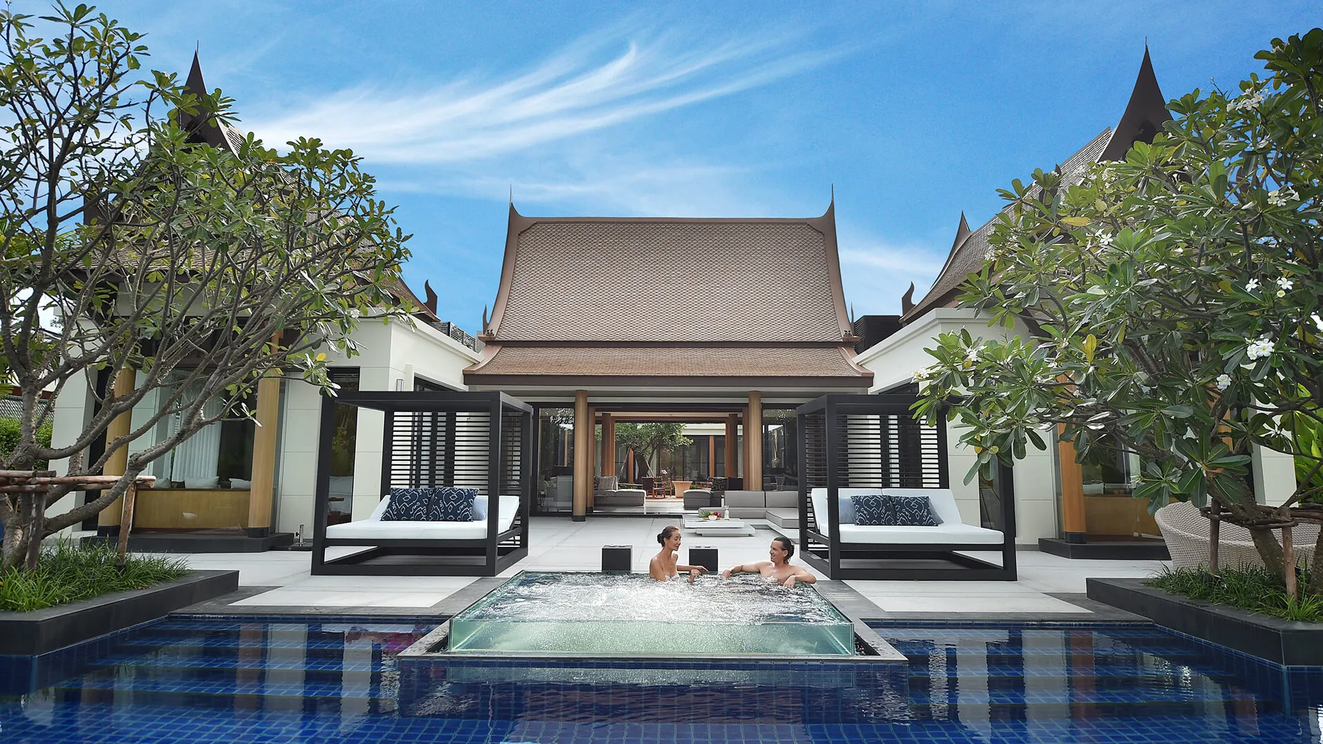 Banyan Tree Grand Residences Phuket - Two individuals are relaxing in a jacuzzi in the foreground, with a scenic traditional-style building and plush landscaping in the background.