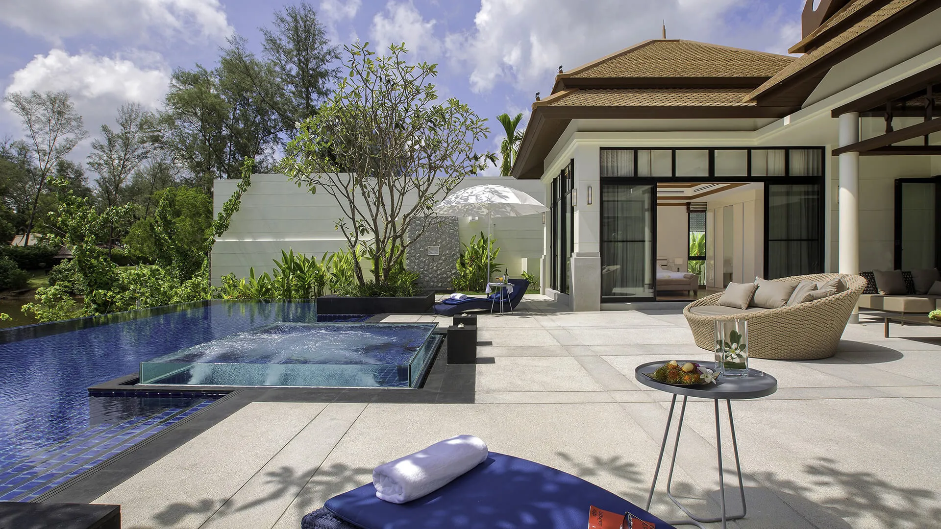 Banyan Tree Grand Residences Phuket - A serene outdoor patio with a blue-tiled pool, surrounded by lush greenery, adjacent to a modern building with large glass doors.