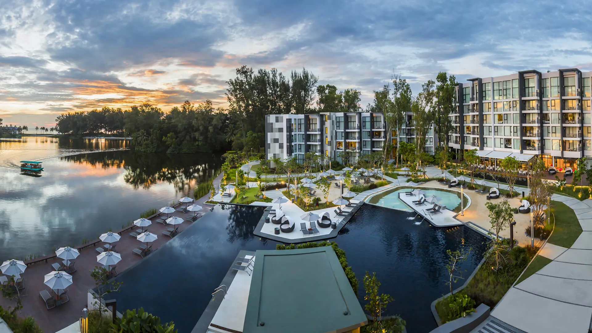 แคสเซีย เรสซิเดนซ์ ภูเก็ต  - Modern resort with swimming pools and loungers, adjacent to a calm river reflecting the sky at sunset.
