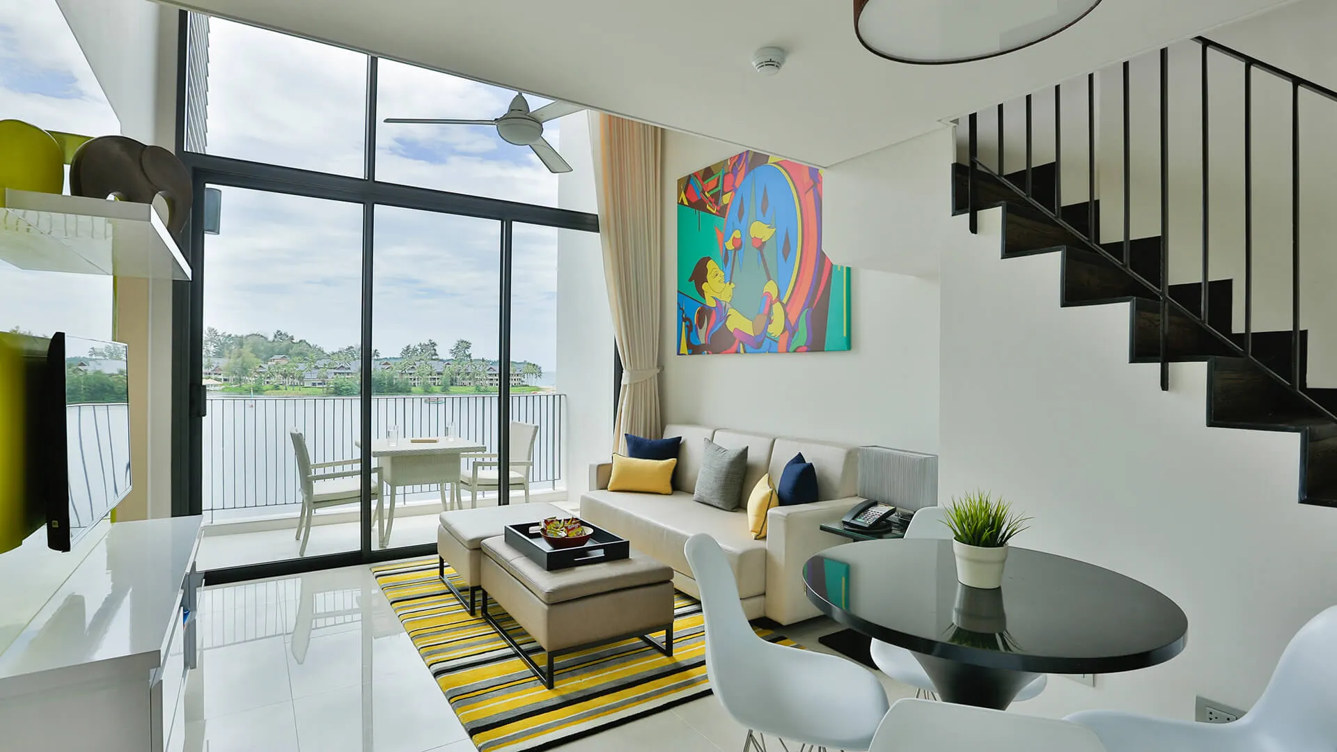 แคสเซีย เรสซิเดนซ์ ภูเก็ต  | A well-lit modern living room with a sofa, a staircase, large windows looking onto a balcony and greenery, and a dining area to the side.