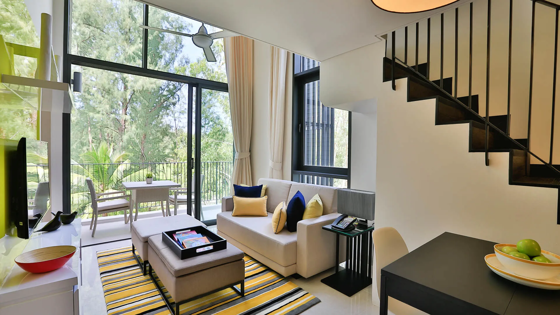 แคสเซีย เรสซิเดนซ์ ภูเก็ต - A well-lit modern living room with a sofa, a staircase, large windows looking onto a balcony and greenery, and a dining area to the side.