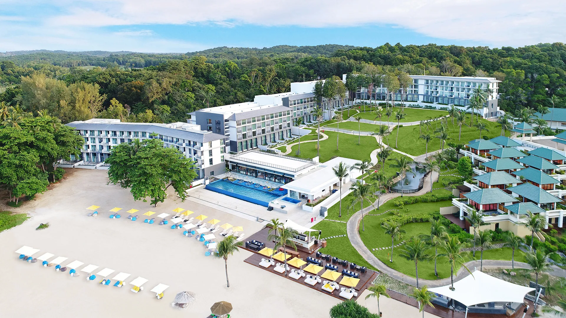 แคสเซีย เรสซิเดนซ์ บินตัน - An aerial view of a coastal resort with modern buildings, a pool by the beach, sun loungers, and surrounding lush greenery.