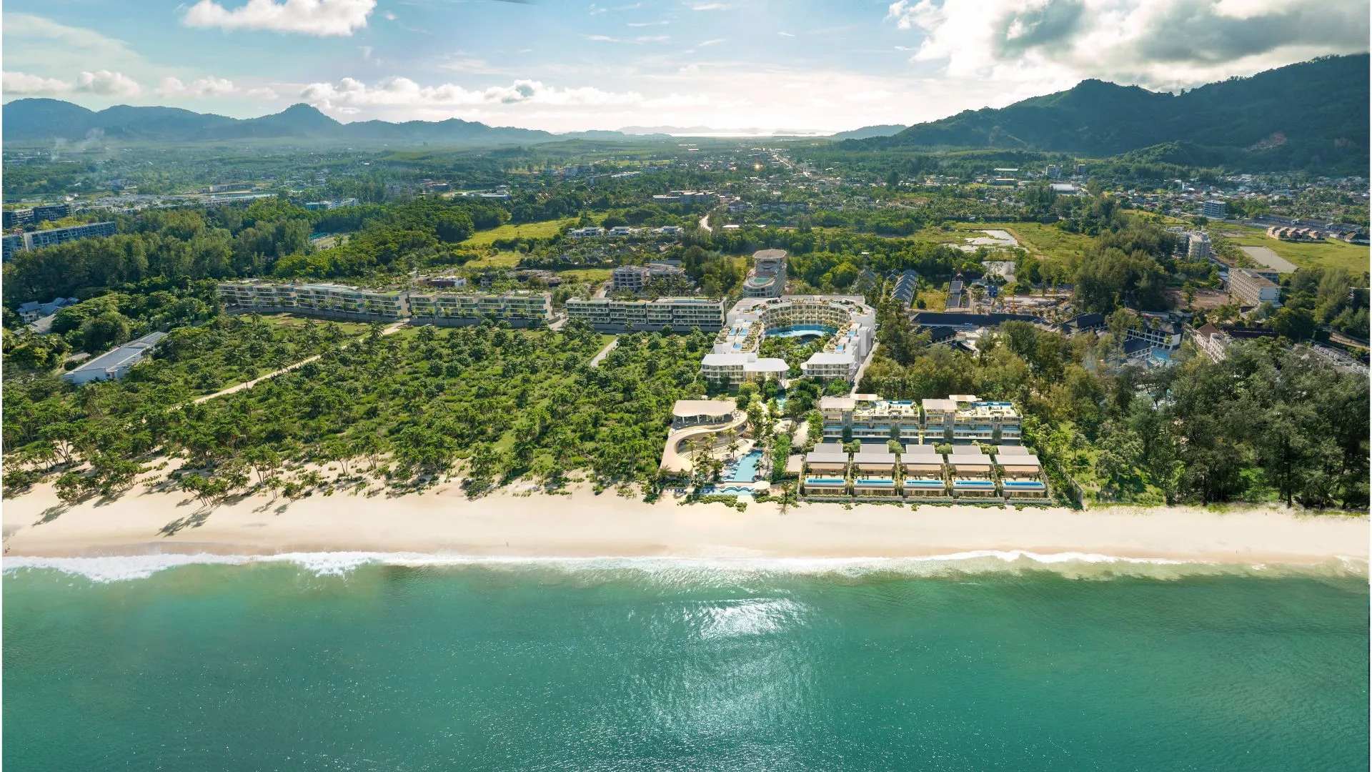 Banyan Tree Beach Residences Varuna | Aerial view of a coastal resort with multiple buildings nestled among greenery beside a sandy beach, with a backdrop of mountains.